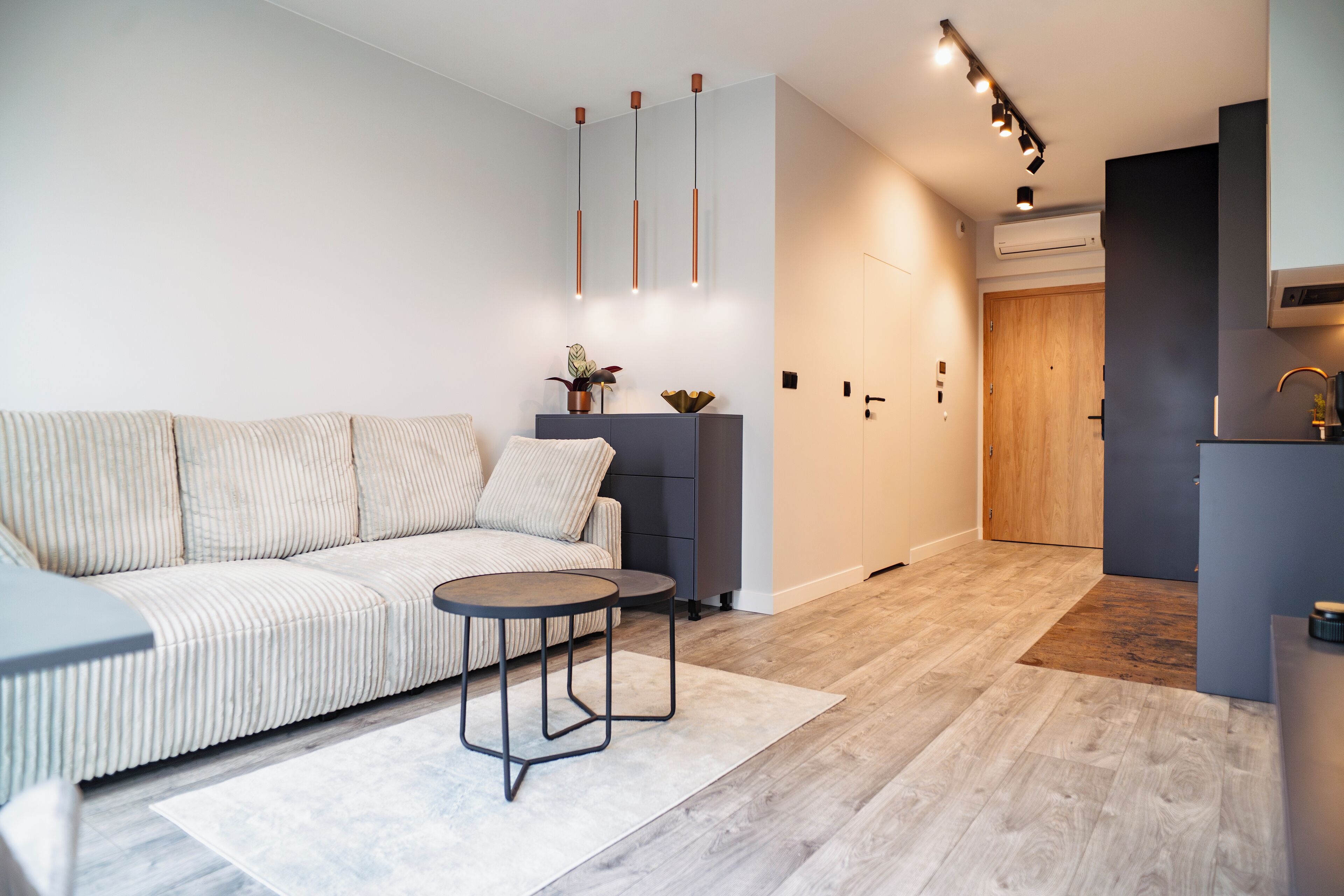 Una sala de estar moderna con un sofá, dos mesitas y una zona de cocina con una puerta de madera y armarios.