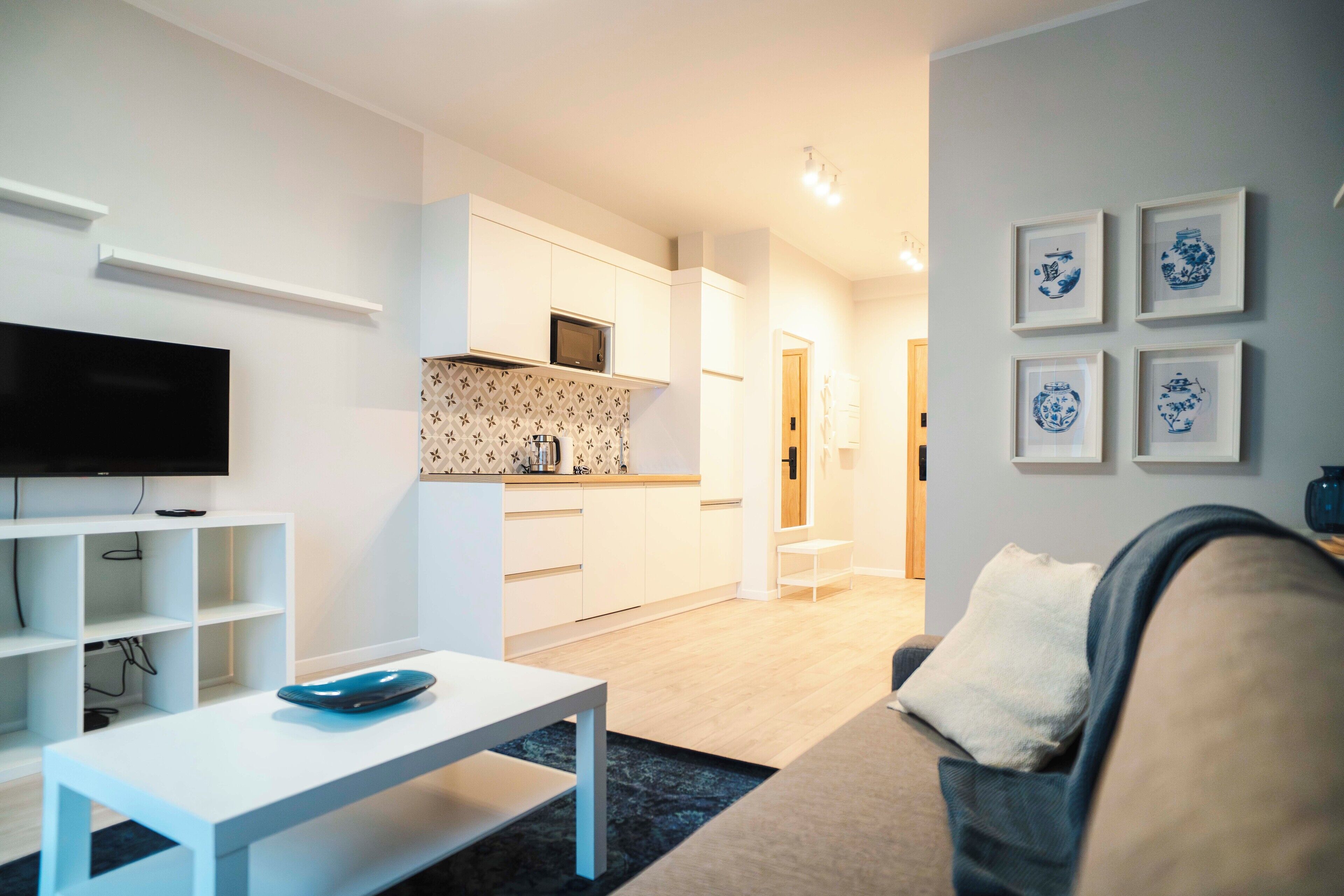 A modern living room with a flat-screen TV, white furniture, a kitchen with white cabinets, and blue-framed artwork on the wall.
