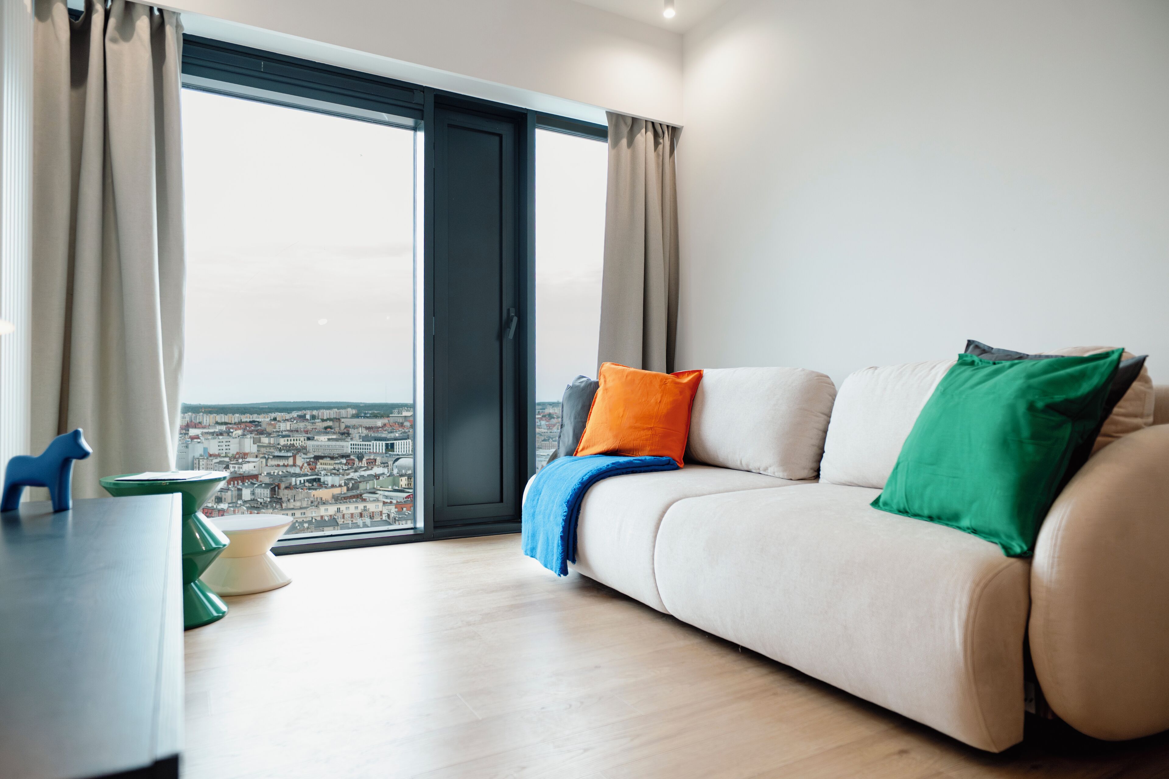 A modern living room with a sofa, a view of a cityscape through large windows, and a table with a blue decorative item.