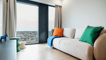 A modern living room with a sofa, a view of a cityscape through large windows, and a table with a blue decorative item.