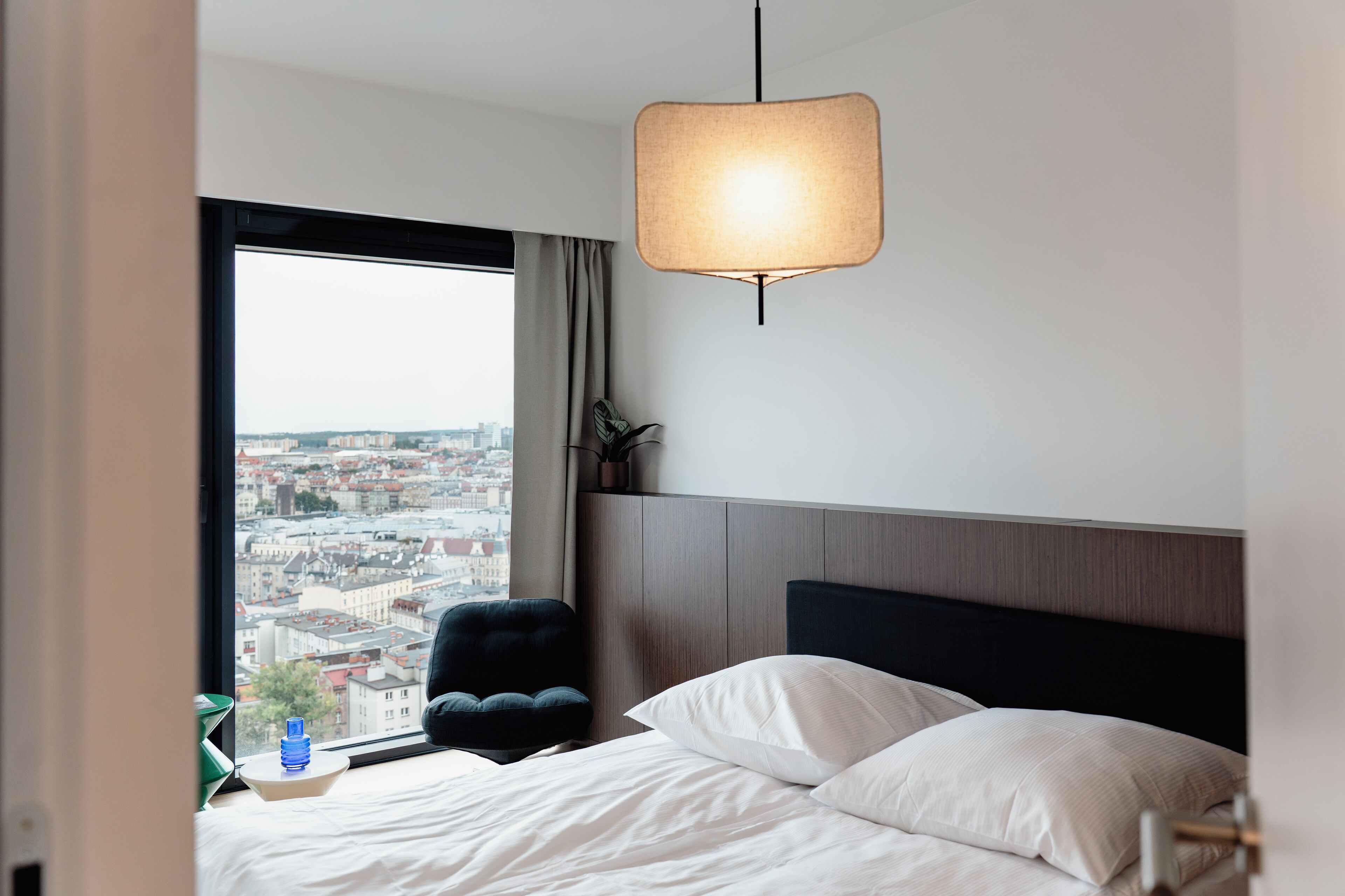 A hotel room with a large window offering a city view, a bed with white linens, a black headboard, and a hanging lamp.