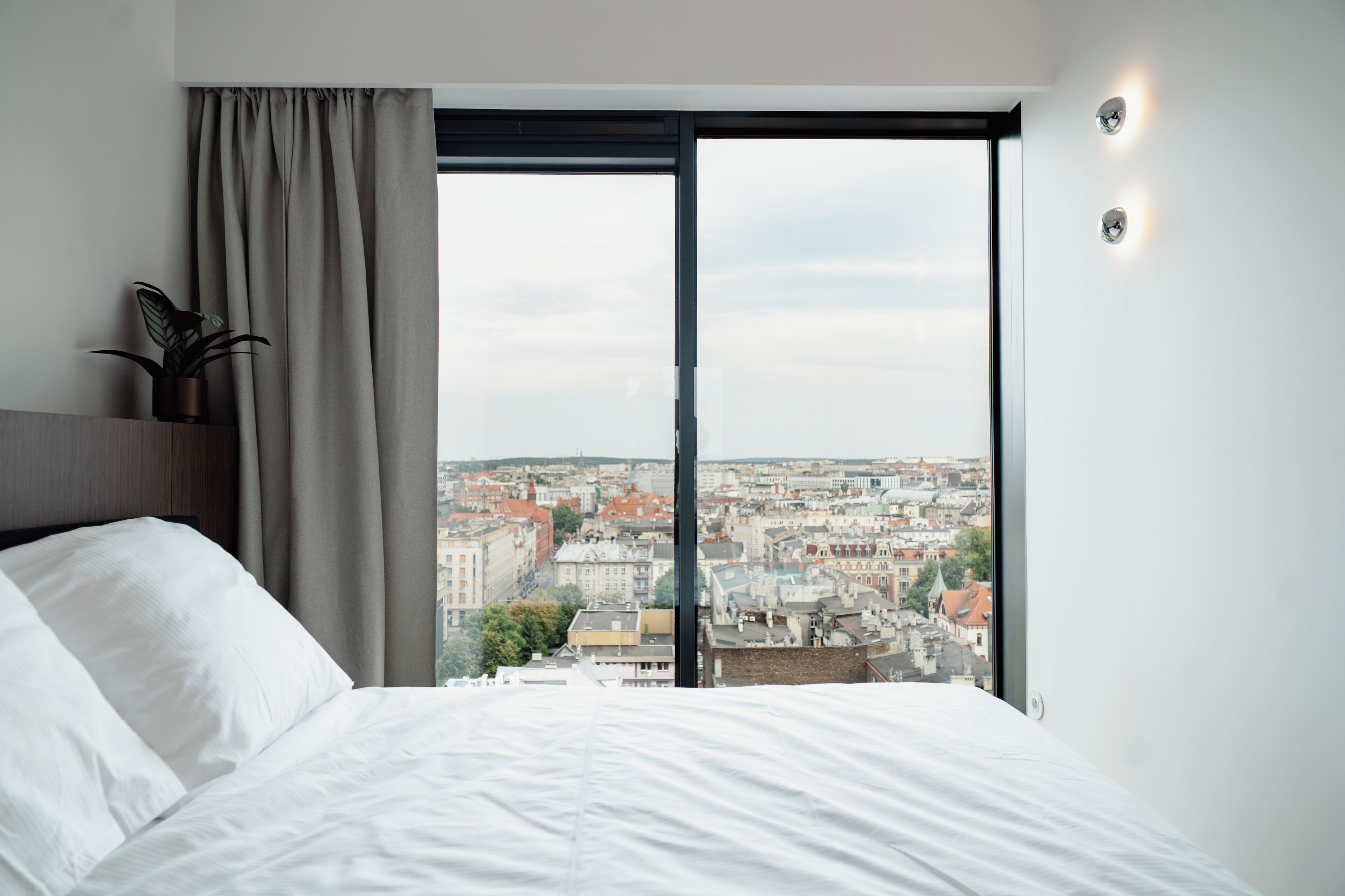 Un dormitorio con una ventana grande, una cama, una planta y vistas a un paisaje urbano.