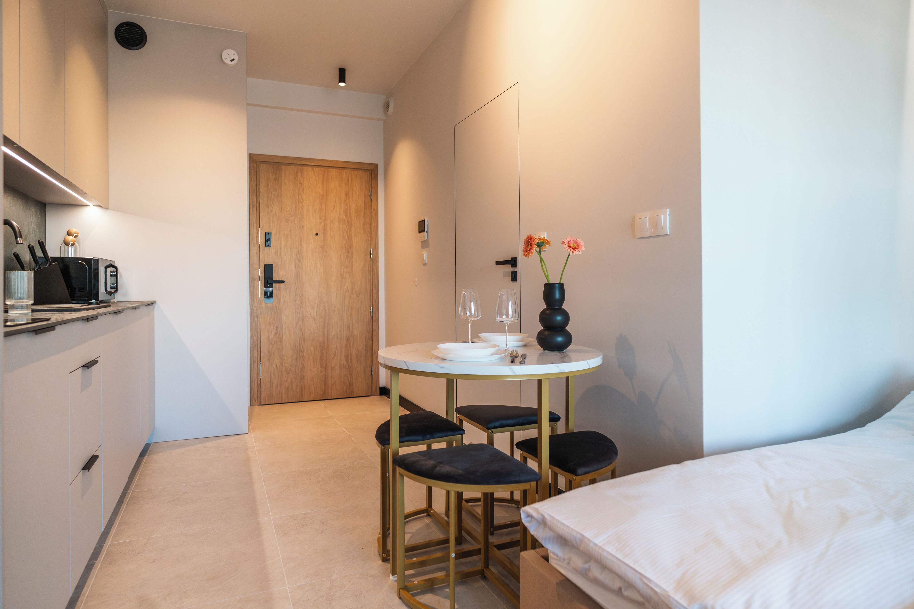 A compact hotel room with a kitchenette, a small dining table with four chairs, a bed, and a vase with flowers.