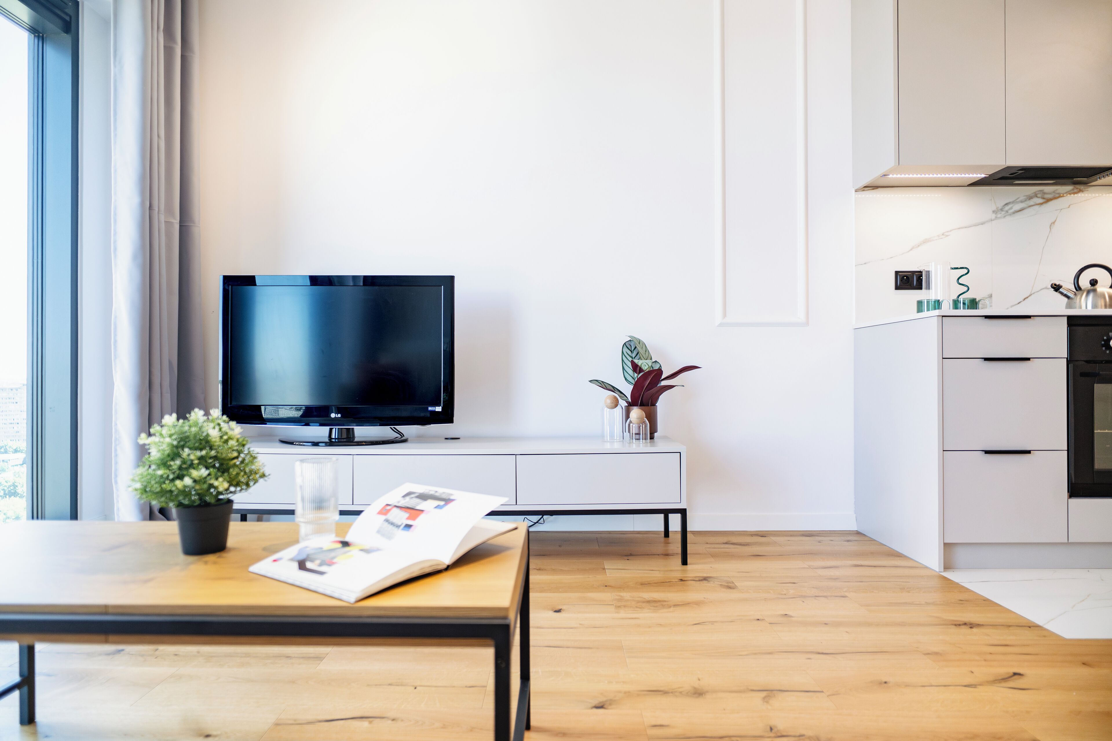 Un salon moderne avec un téléviseur à écran plat, une table basse en bois sur laquelle repose un magazine, une plante en pot et un meuble blanc contenant des appareils de cuisine.