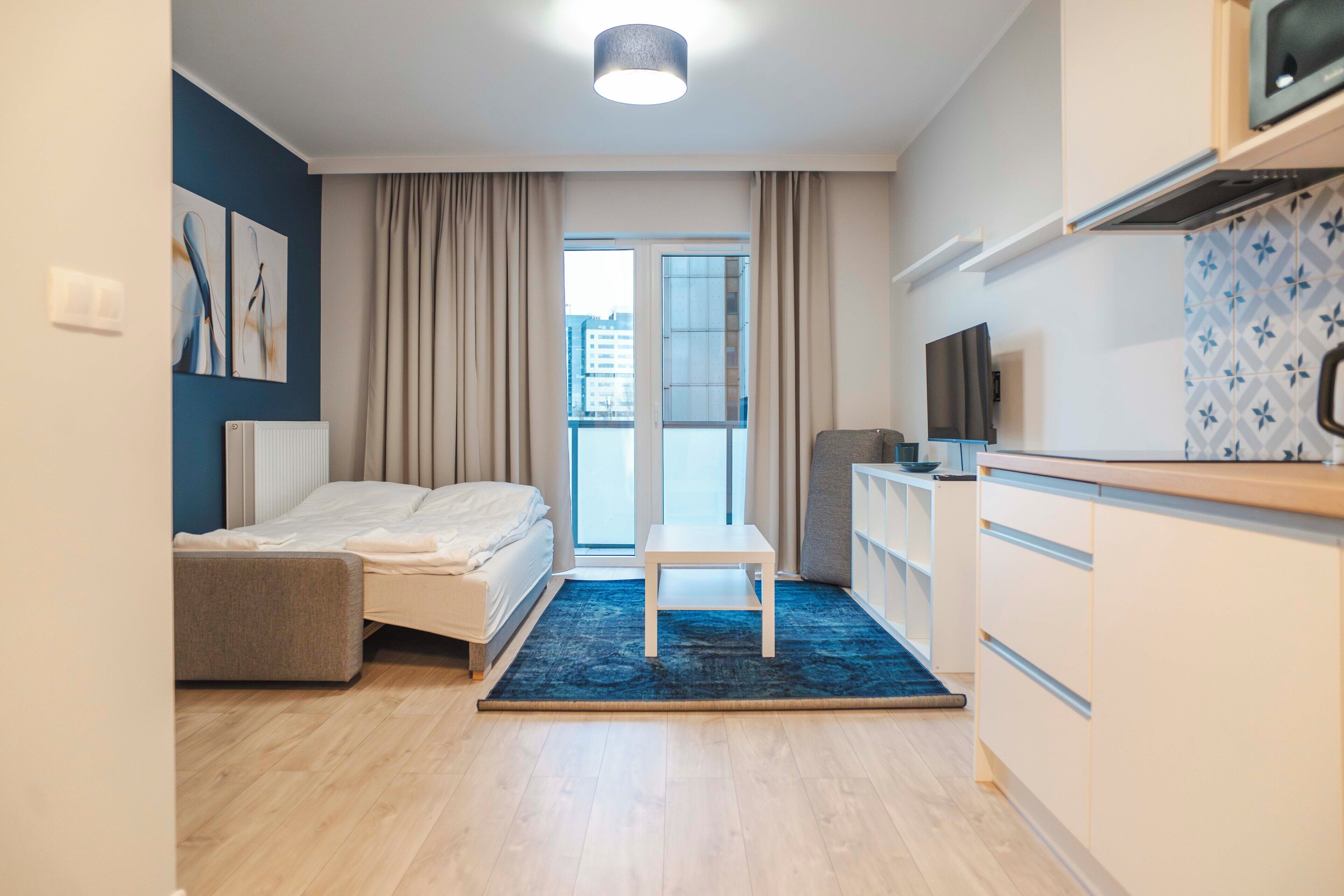Een moderne hotelkamer met een bed, een klein tafeltje, een kitchenette en uitzicht op de stad door het raam.