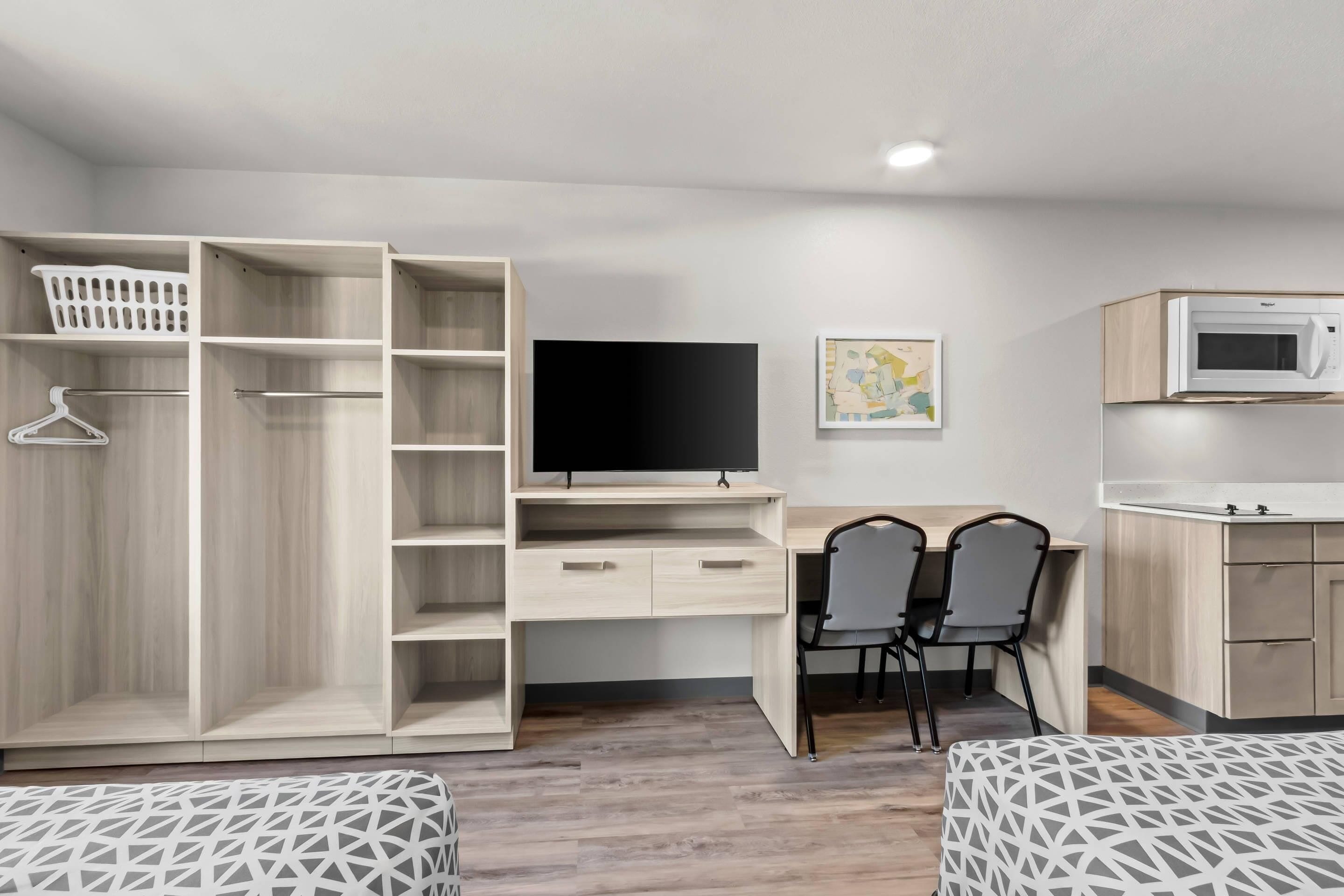 Une chambre moderne équipée d’une armoire en bois, d’une télévision, d’un bureau avec deux chaises, d’un four à micro-ondes et d’un lit avec une literie à motifs.