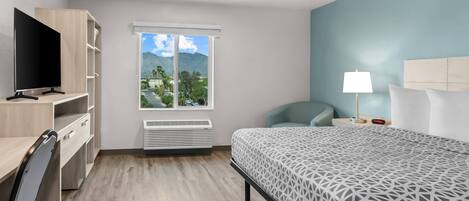 A hotel room with a bed, a desk with a TV, a chair, a window with a view of mountains, and a lamp.