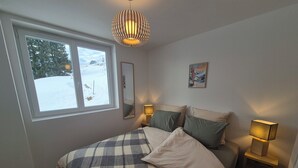 Ein Schlafzimmer mit großem Fenster, einem Bett mit kariertem Überwurf, zwei Nachttischlampen, einem Spiegel und einem Bild an der Wand.