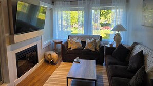 A living room with a flat-screen TV, a fireplace, a sofa, a coffee table, and large windows with curtains. - Oceanside Gardens One Bedroom Ground Floor w/ Kitchen at Pacific Shores Resort! (Nanoose Bay)