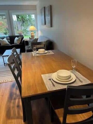 Un coin repas avec une table en bois dressée pour deux personnes, un canapé en arrière-plan et une fenêtre donnant sur un paysage verdoyant.