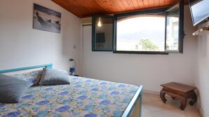 A bedroom with a bed, a small wooden table, a window with an open shutter, and a painting on the wall.