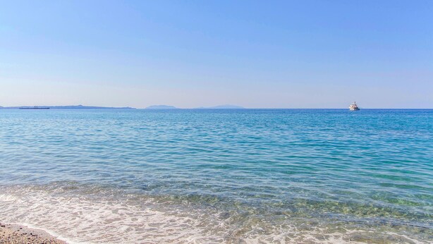 A calm sea with clear water near the shore, a distant ship, and a small island in the background.