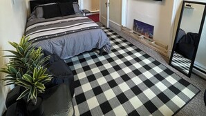 A bedroom with a checkered floor, a bed with striped bedding, a TV mounted on the wall, and a plant on the left.