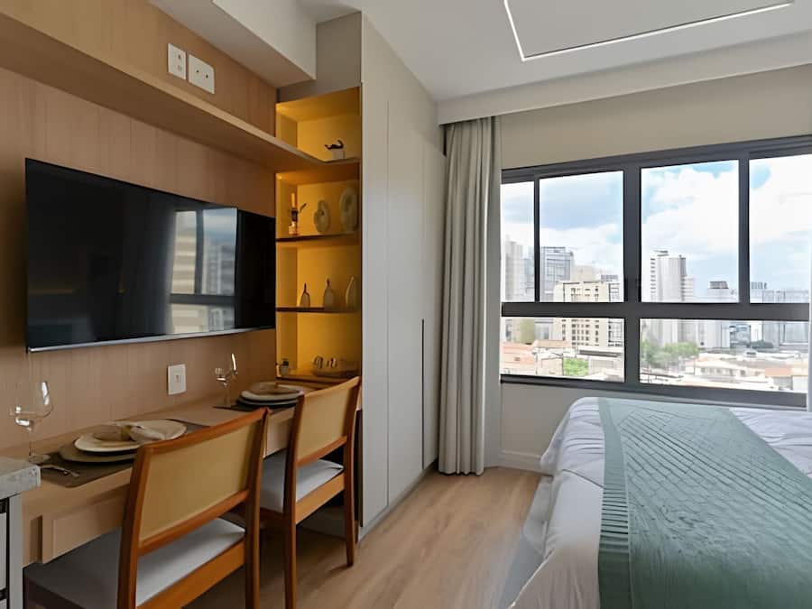 Quarto de hotel com cama, escrivaninha com cadeiras, televisão de tela plana e uma grande janela com vista para a cidade.