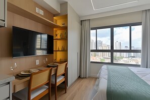 A hotel room with a bed, a desk with chairs, a flat-screen TV, and a large window with a city view. - Prime Smart Santa Cruz (São Paulo)