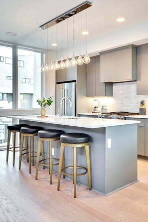 A modern kitchen with a large island, bar stools, stainless steel appliances, and a view of the city. - Modern Bachelorette Retreat — Rooftop, Karaoke, Near Downtown Broadway. (Nashville)