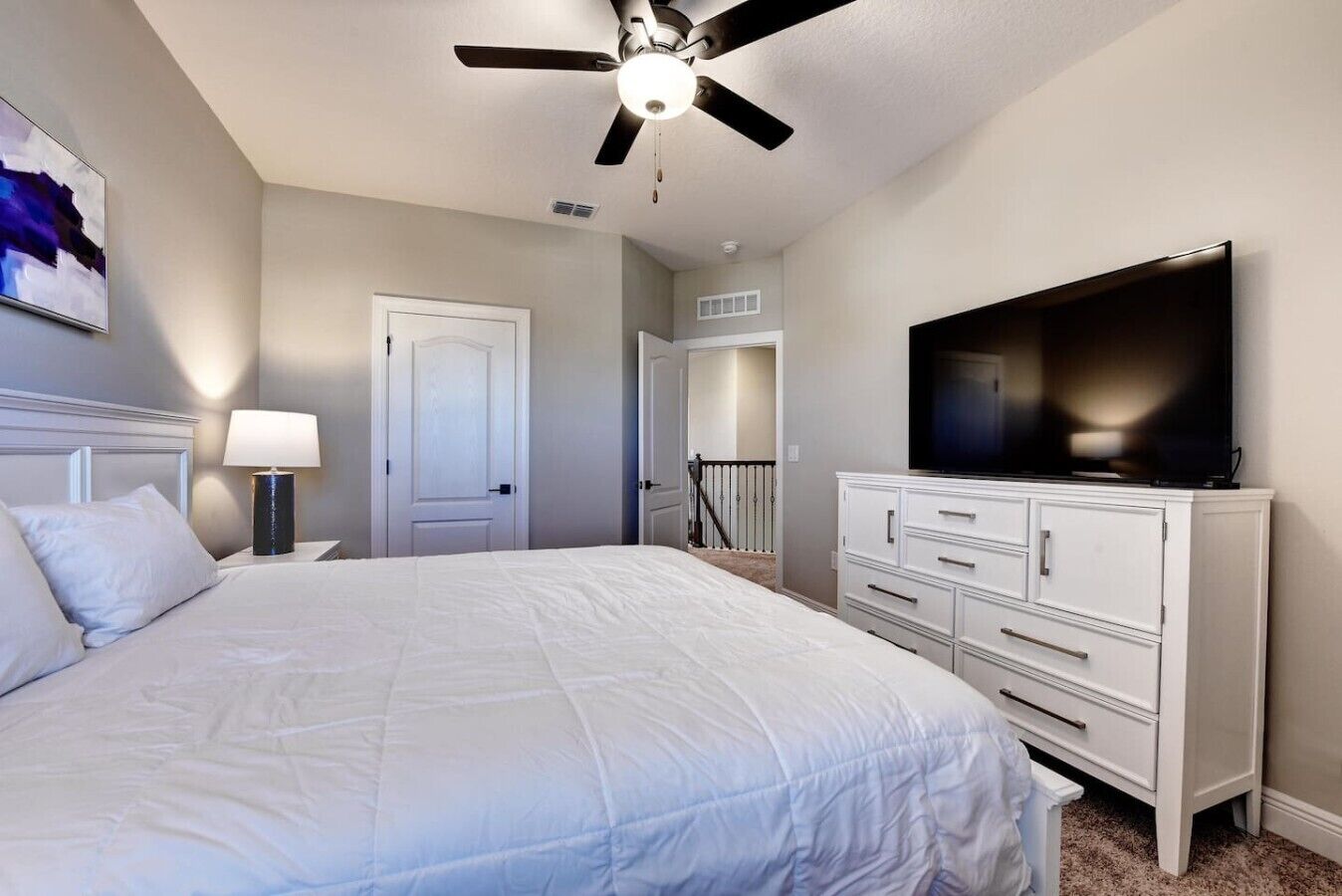 Une chambre à coucher avec un lit, un ventilateur de plafond, une télévision, une commode et une porte.