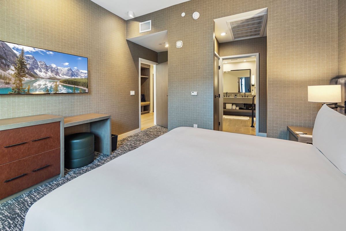 A hotel room with a large bed, a desk with drawers, a trash bin, a bathroom with a mirror, and a wall-mounted TV displaying a mountain landscape.