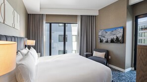 Habitación de hotel con una cama grande, un escritorio con lámpara, una silla, una ventana con cortinas y un cuadro de un paisaje montañoso en la pared.