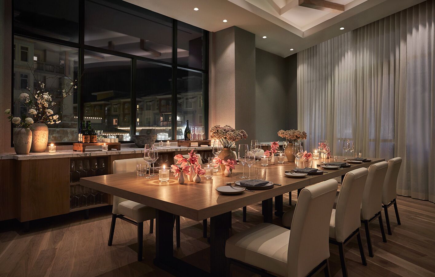 A modern dining area with a long table set for six, featuring a mix of floral centerpieces and candles.