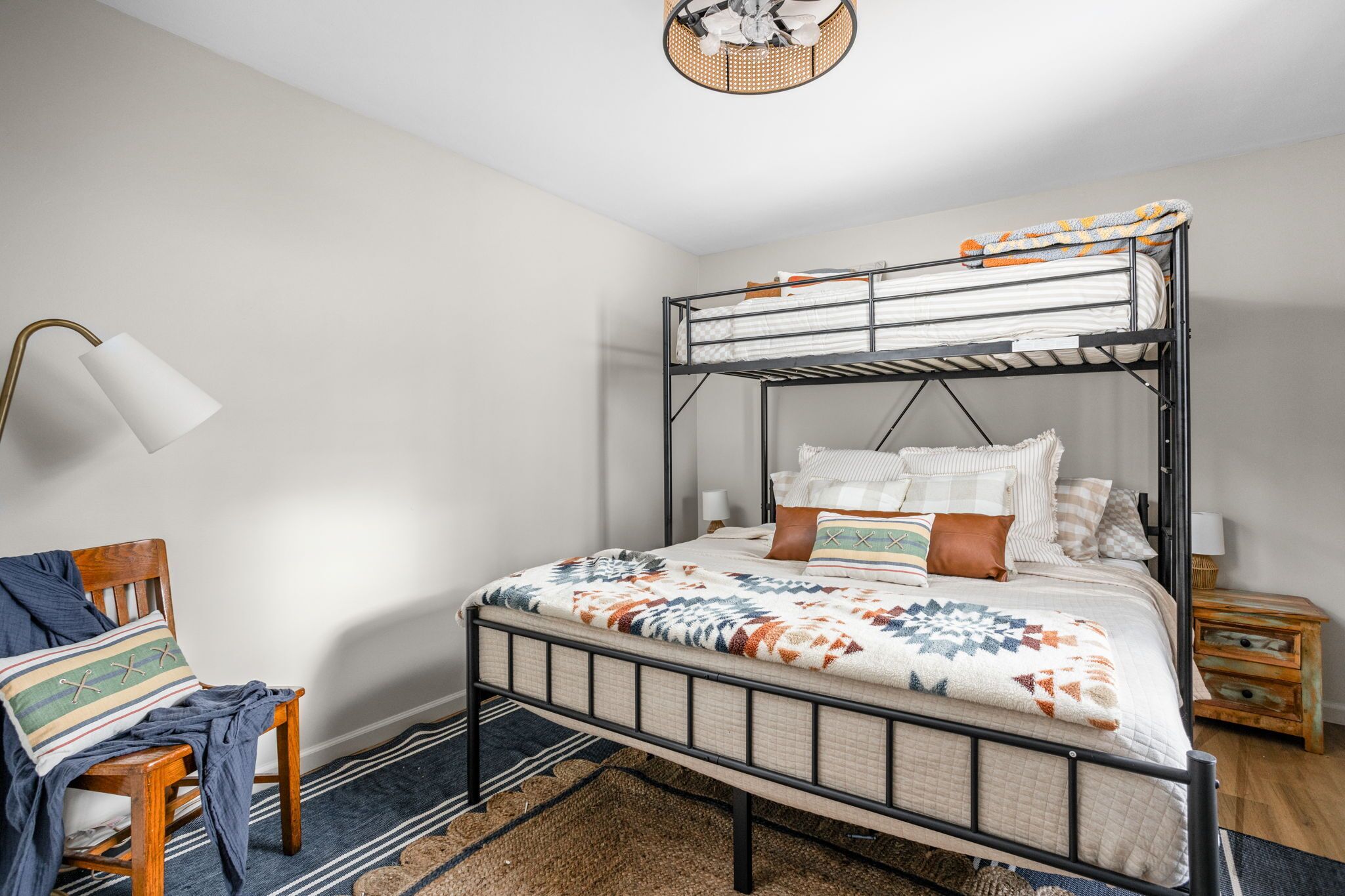 A bunk bed with a patterned blanket, a wooden chair with a blanket, a lamp, and a small wooden nightstand.