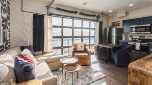 Una sala moderna con sofá, sillón y mesa de centro, una cocina con electrodomésticos y ventanas amplias con vistas a la ciudad.