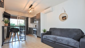 A modern apartment with a grey sofa, a dining area with a table and chairs, a kitchen with an oven, and a balcony with a view.