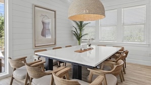 Uma sala de jantar com uma mesa branca, cadeiras de madeira, uma luminária pendente trançada, uma obra de arte emoldurada e um vaso com planta.