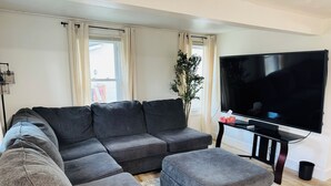 A modern living room with a sectional sofa, a flat-screen TV, and a small table. - Super Modern 2-Bedroom Riverside Home @ RIT (Rochester)