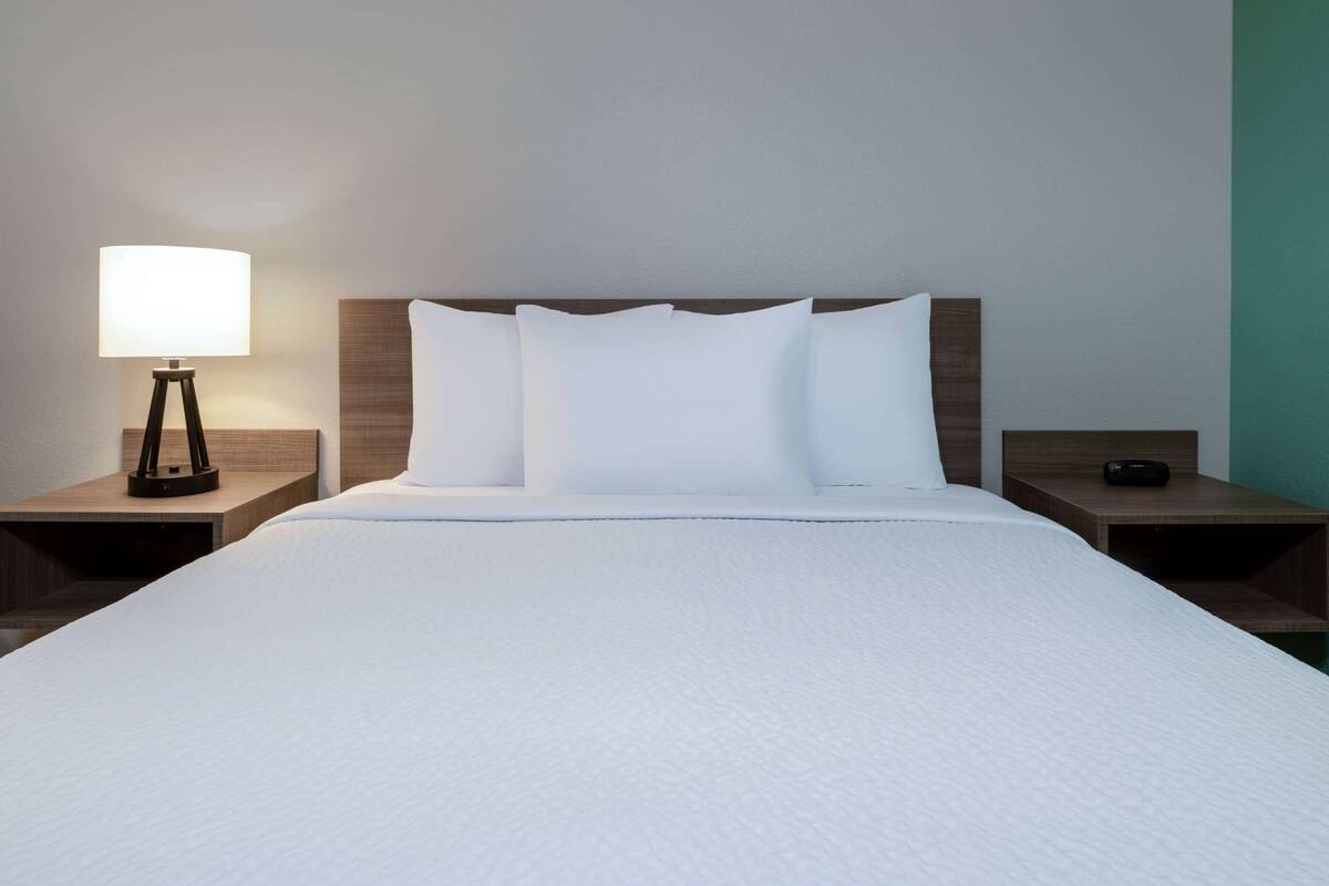 A neatly made bed with white linens and pillows, a bedside lamp, and a wooden nightstand.