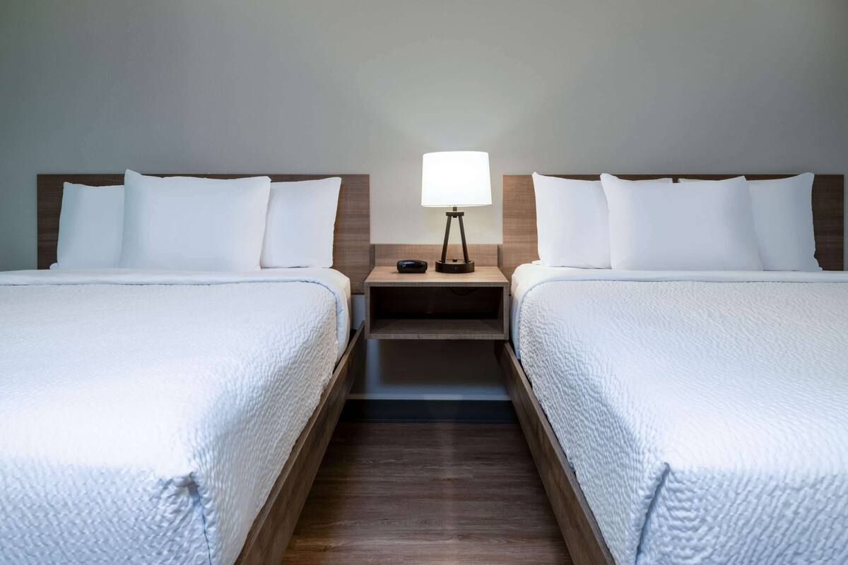 A hotel room with two beds, a nightstand with a lamp, and a small shelf.