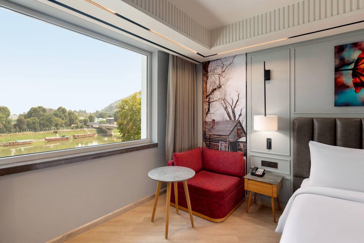 A hotel room with a large window, a red sofa, a small round table, a bedside lamp, and a bed.