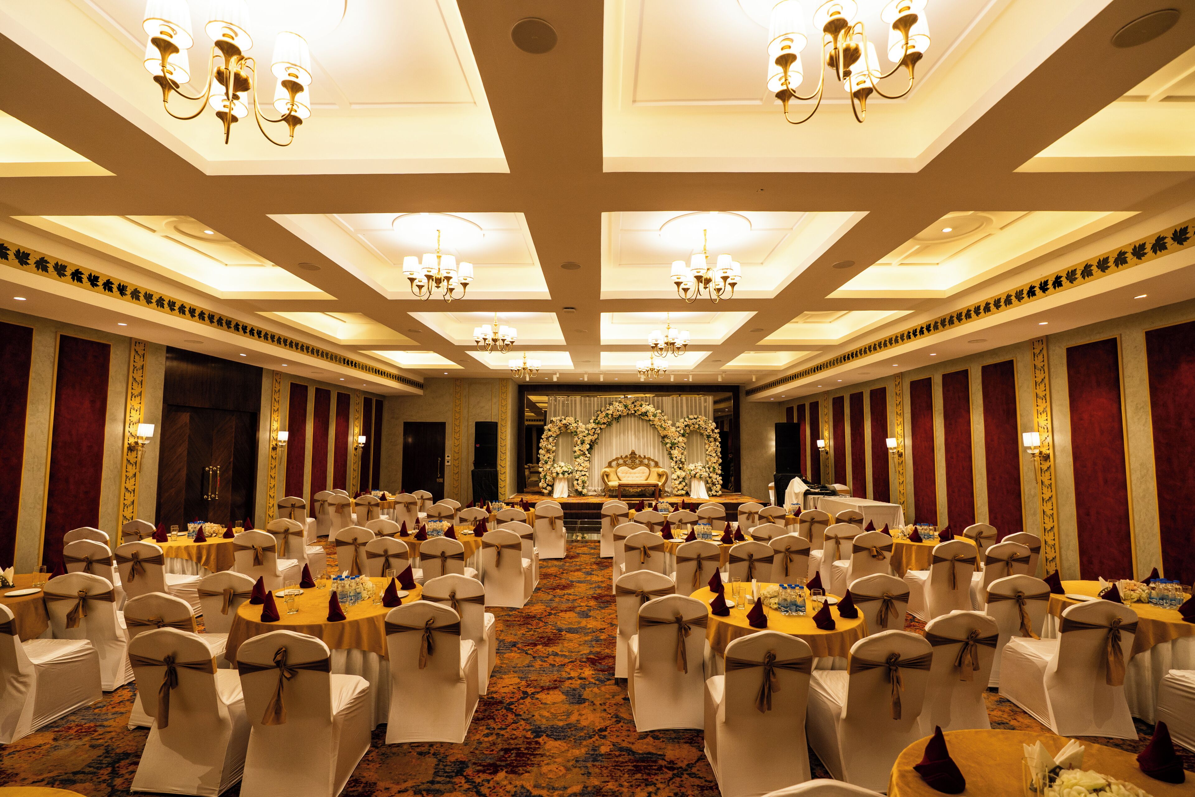 A banquet hall with round tables, white chairs, chandeliers, and a stage with floral decorations.