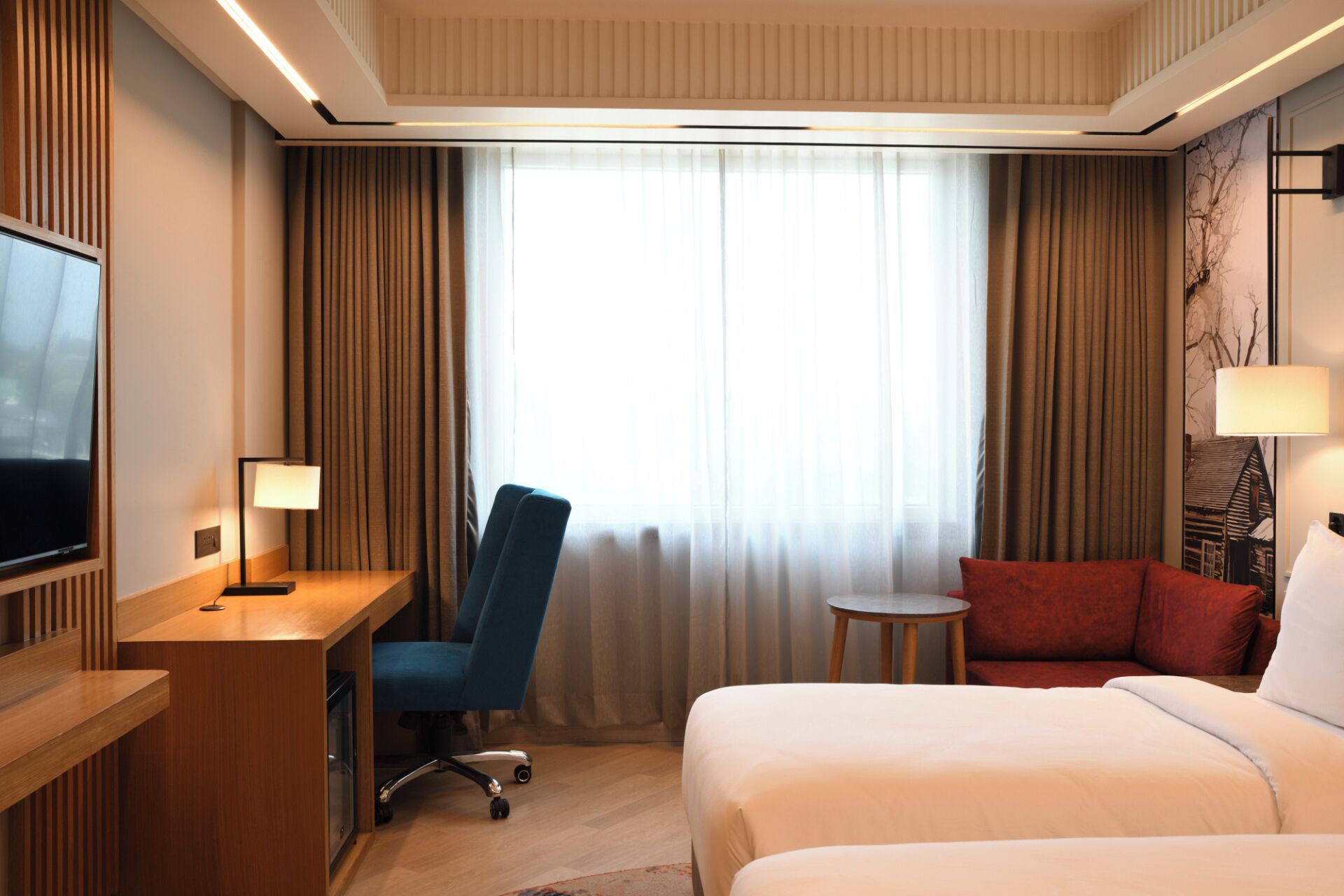 A hotel room with a bed, a desk with a chair, a TV, a small table, and a window with curtains.