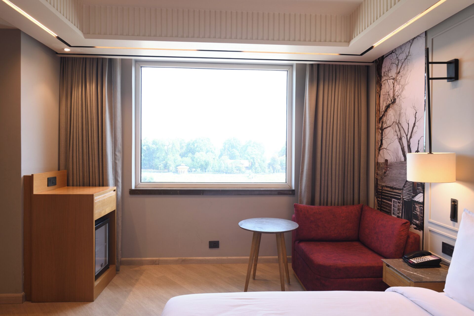 Habitación de hotel con cama, sofá rojo, una mesita redonda, lámpara, teléfono y ventana con cortinas.