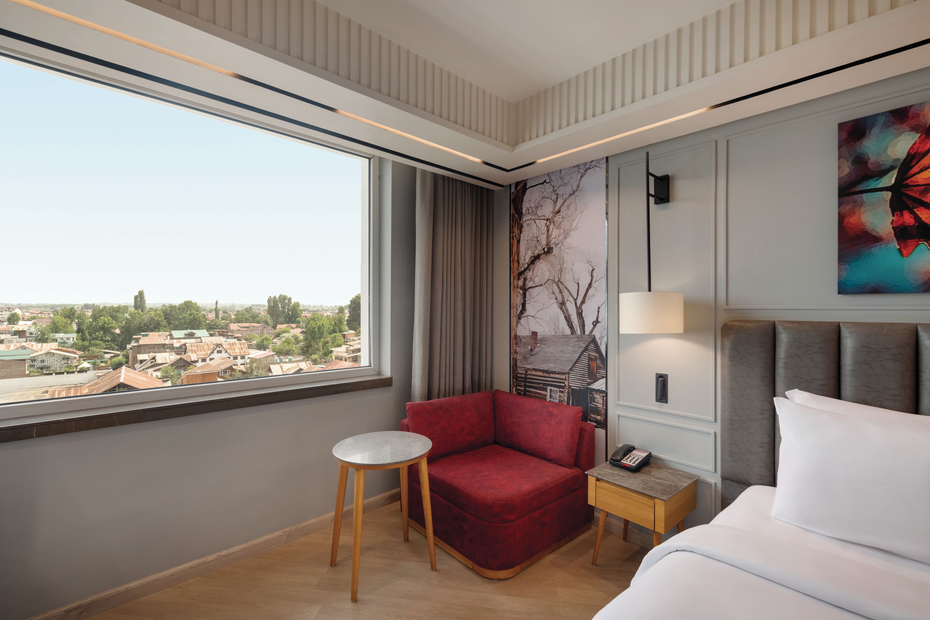 Quarto de hotel com uma janela grande, um sofá vermelho, uma mesinha redonda, uma mesa de cabeceira com abajur e uma cama com roupa de cama branca.