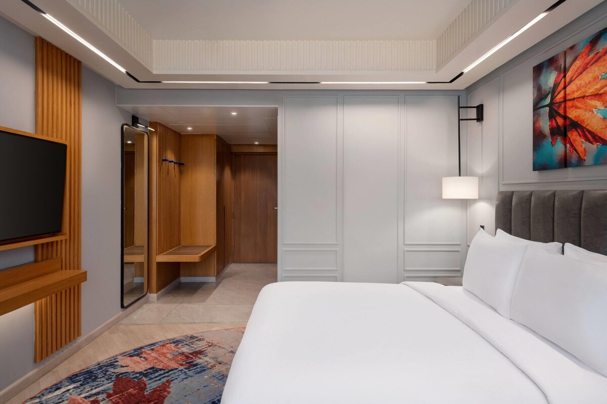 A modern hotel room with a large bed, a TV mounted on the wall, a walk-in closet, and a colorful abstract painting on the wall.