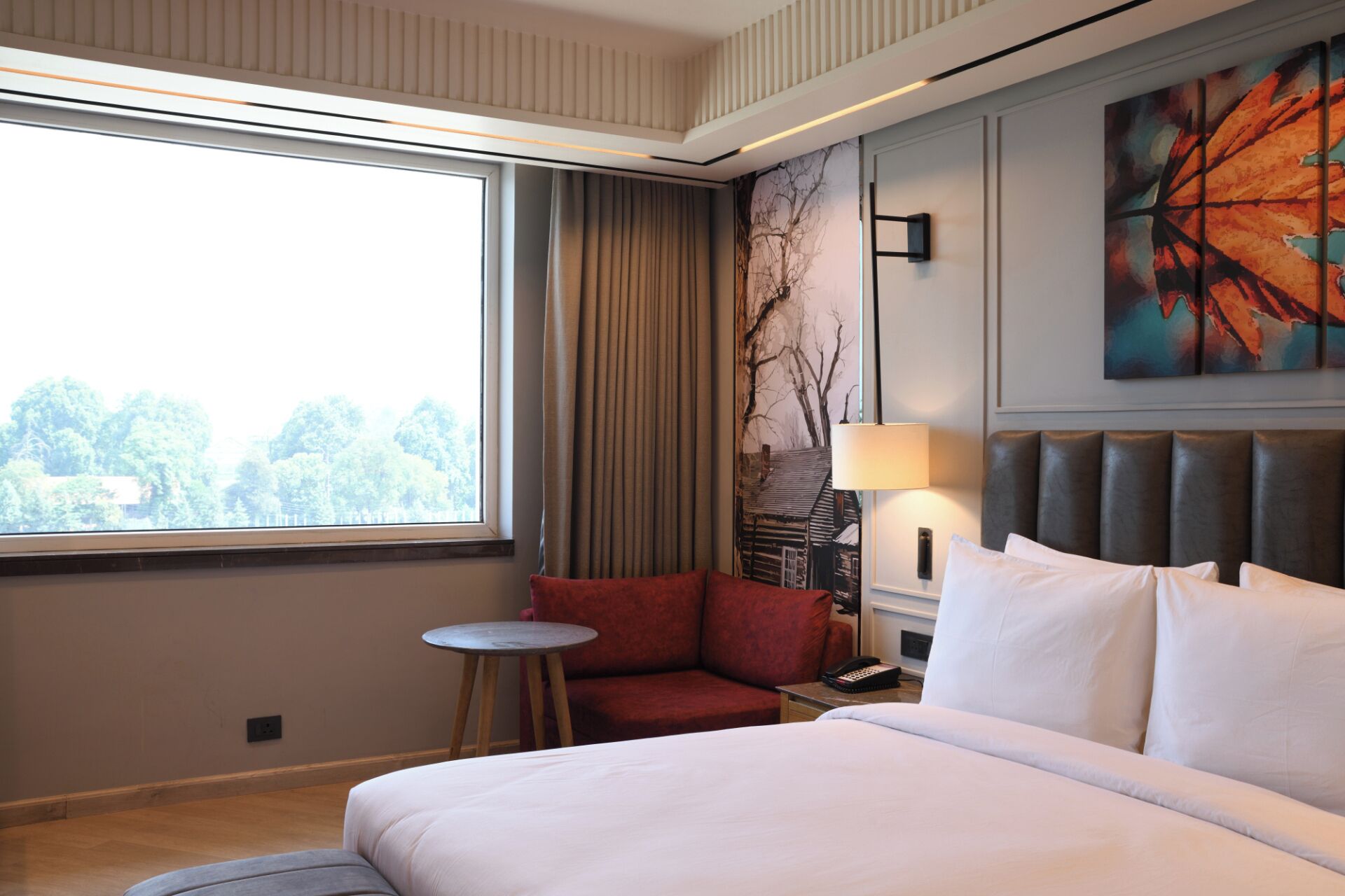 Habitación de hotel con una cama grande, una mesita, una silla roja, una lámpara y un cuadro en la pared.