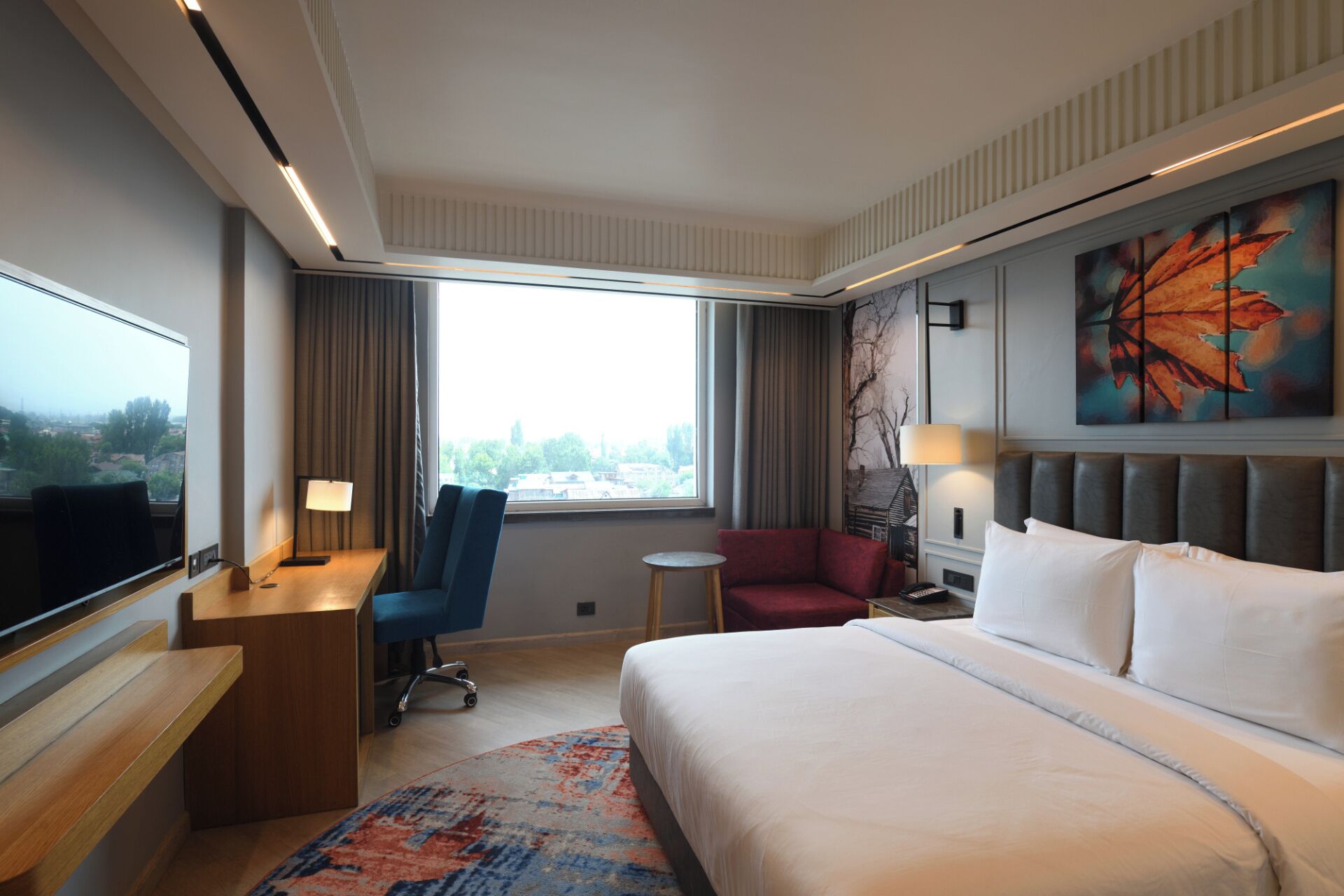 A hotel room with a bed, a desk, a chair, a TV, a window with curtains, and a painting on the wall.