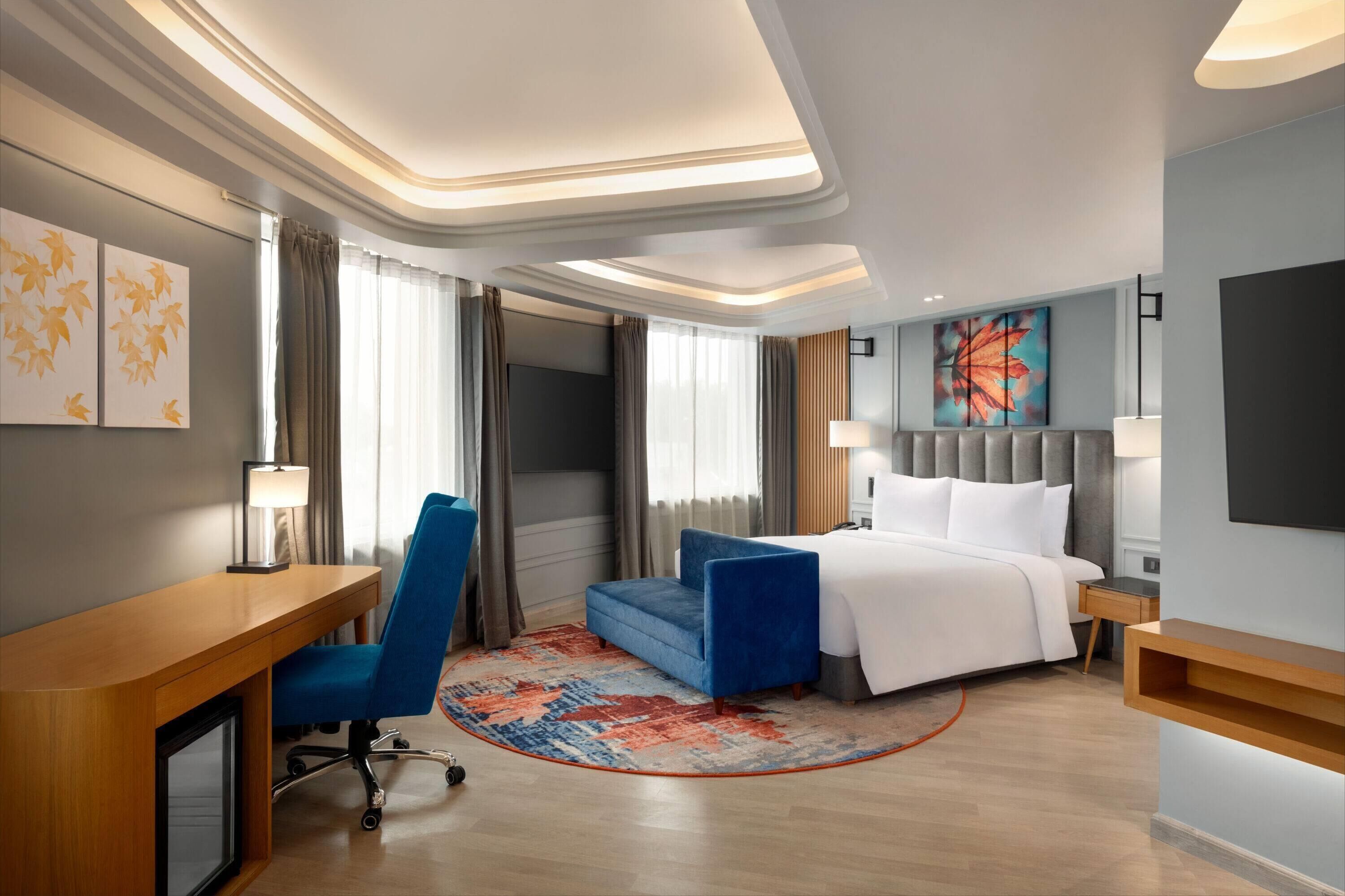 Quarto de hotel moderno com cama, escrivaninha, cadeira e uma janela grande com cortinas.