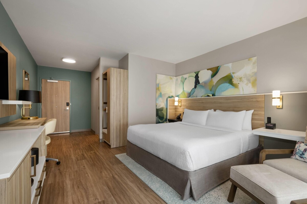 A hotel room with a bed, desk, chair, and a large abstract painting on the wall.