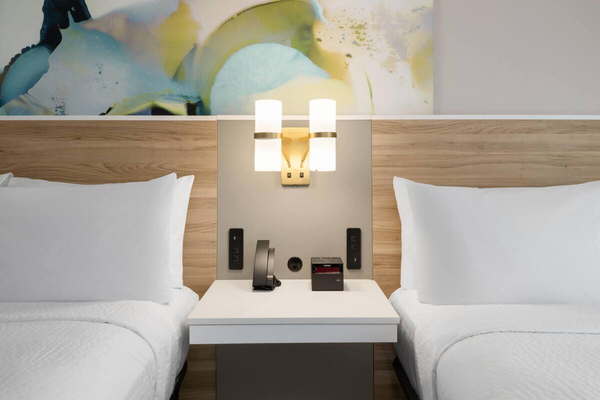 Une chambre d’hôtel avec deux lits, une table de chevet avec une lampe, un téléphone et une petite horloge.