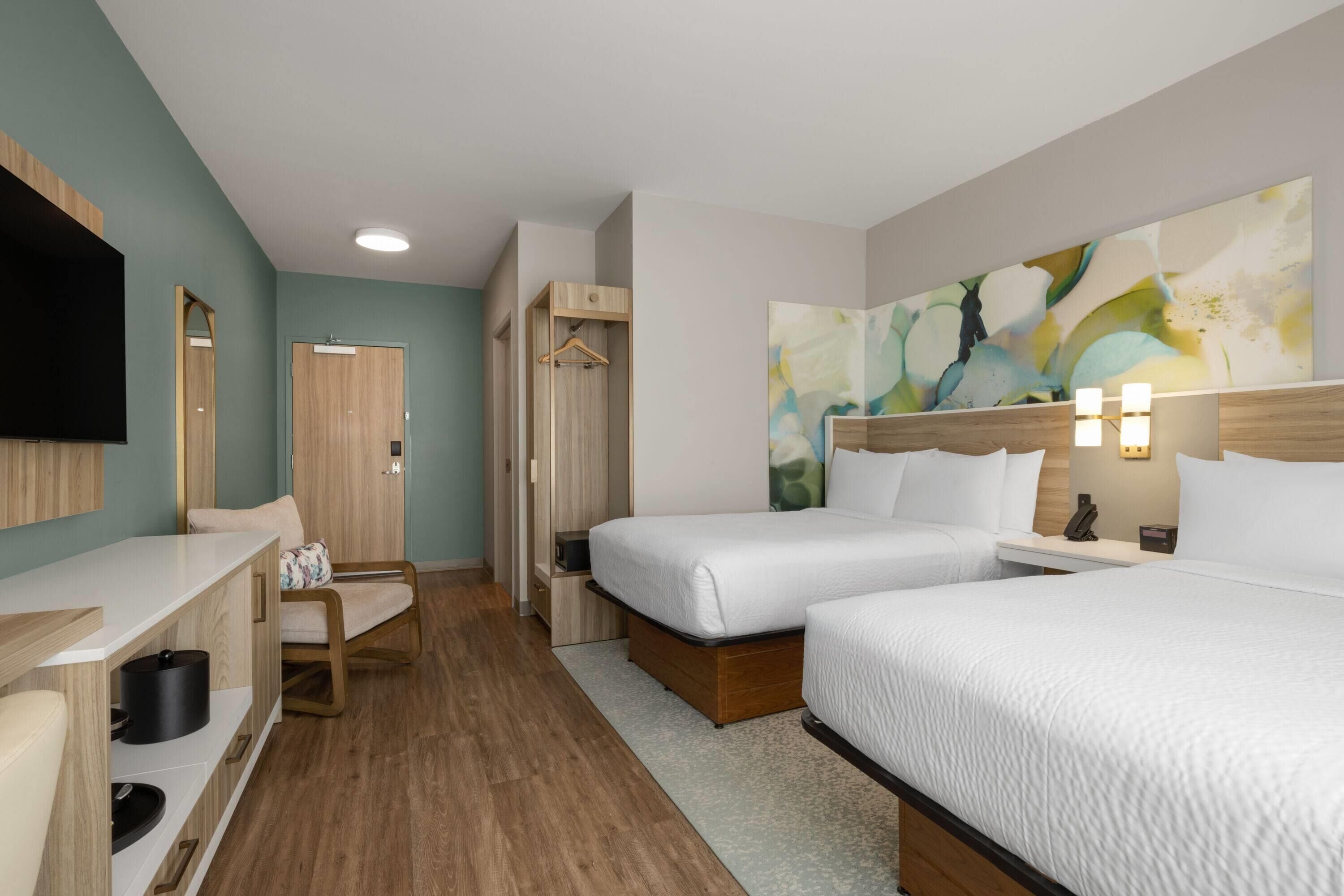 Habitación de hotel con dos camas, un escritorio, una silla, un televisor y un cuadro en la pared.