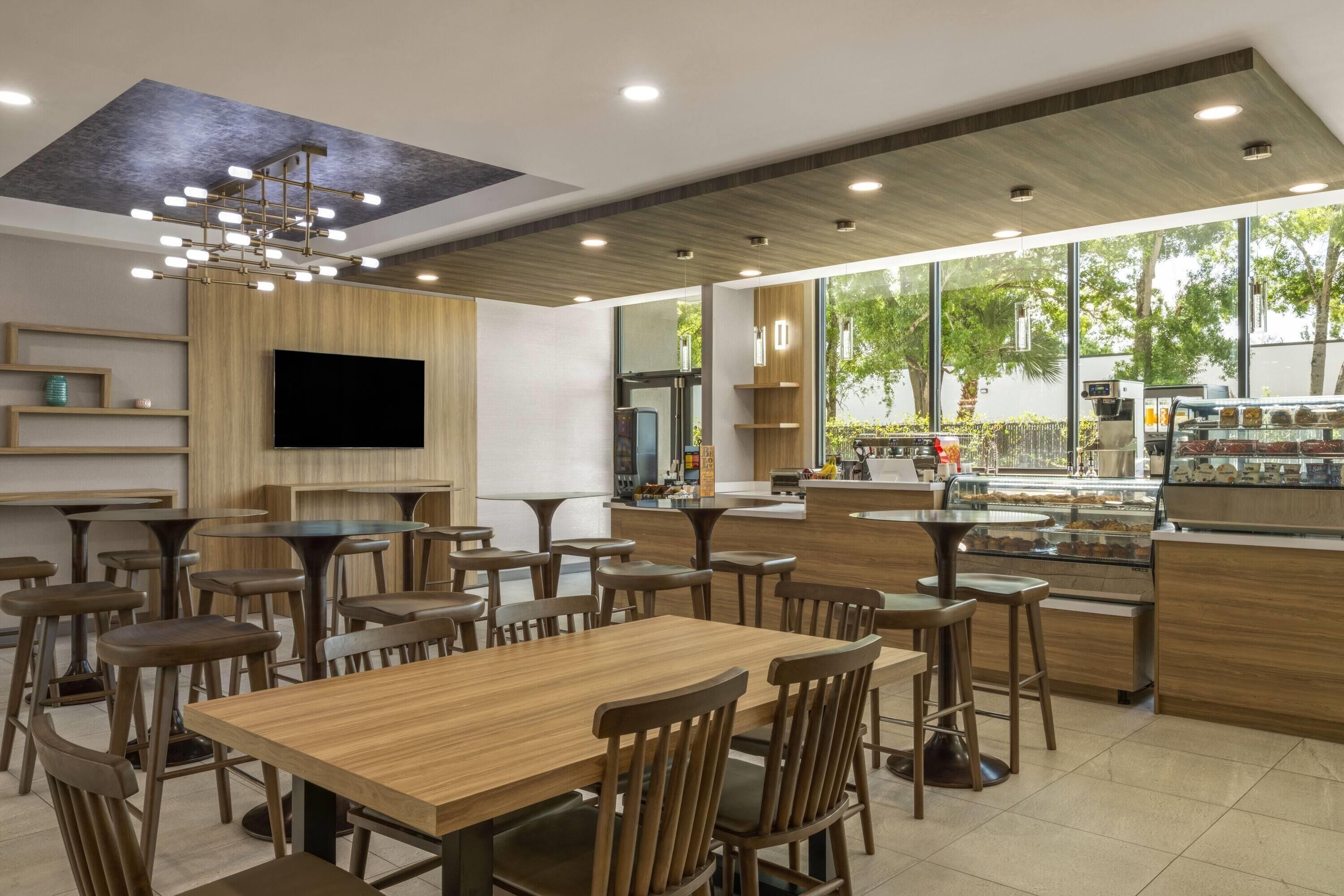 A modern restaurant with wooden tables and chairs, a flat-screen TV, and a large window offering a view of the outdoors.