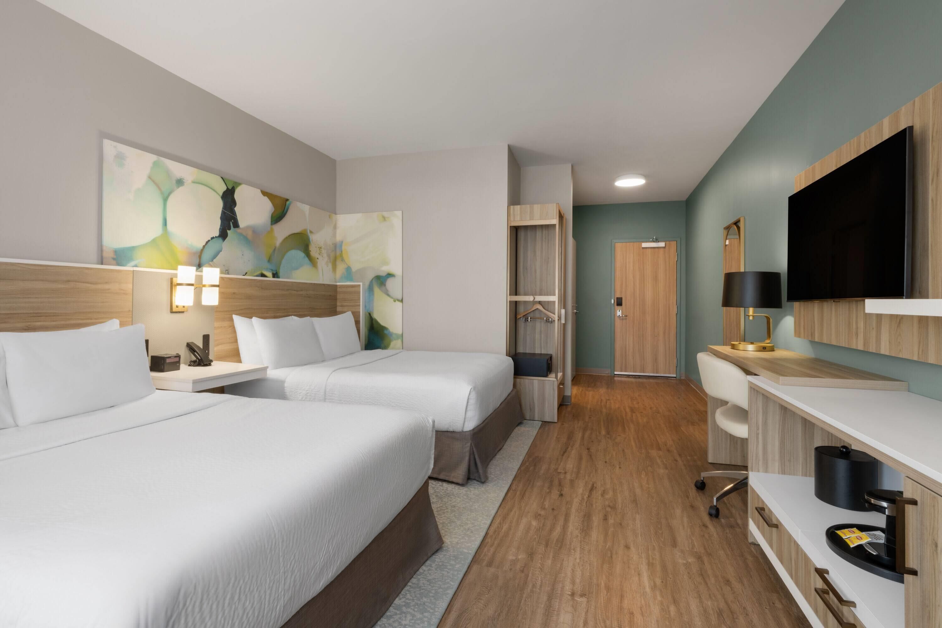 A hotel room with two beds, a TV, a desk, a chair, and a painting on the wall.