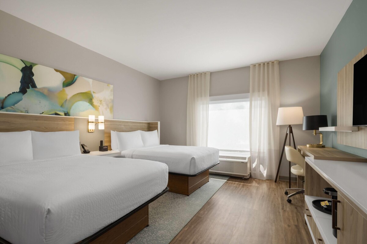 A hotel room with two beds, a desk, a chair, a lamp, a TV, and a large abstract painting on the wall.