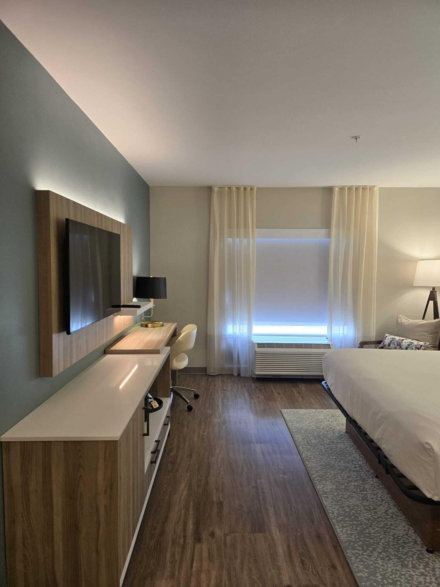 A modern hotel room with a wooden desk, a bed, a large window with curtains, and an air conditioning unit.