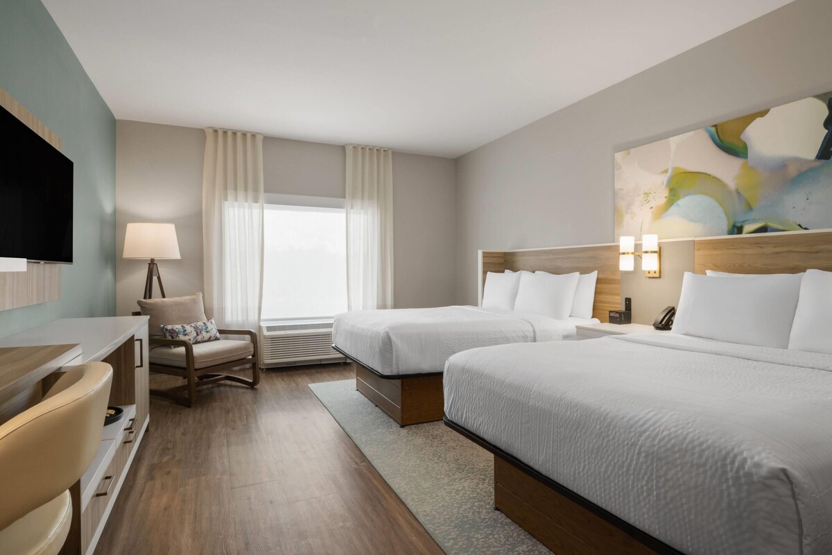 A hotel room with two beds, a desk, a chair, a TV, and a painting on the wall.