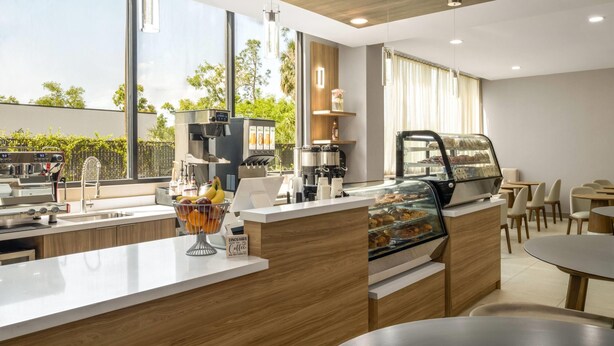 A modern coffee shop with a counter, coffee machines, a display case with pastries, and a view of greenery outside. - Wyndham Garden Tampa Fairgrounds/Casino (Tampa)