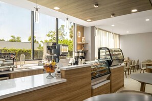 A modern coffee shop with a counter, coffee machines, a display case with pastries, and a view of greenery outside. - Wyndham Garden Tampa Fairgrounds/Casino (Tampa)